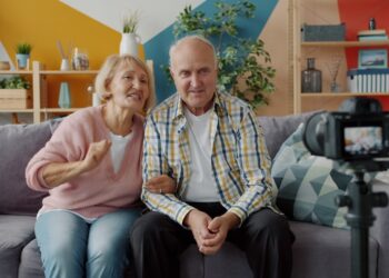 Senior bloggers man and woman are recording video showing thumbs-up using camera at home making content for internet vlog. People and technology concept.