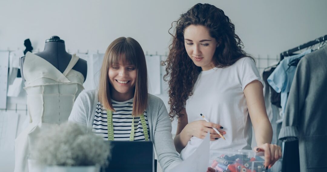 Cheerful creative clothing designers are watching laptop screen, having friendly conversation, gesturing, smiling and laughing together in light workshop. Fun at workplace concept.