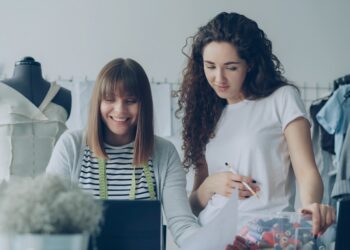 Cheerful creative clothing designers are watching laptop screen, having friendly conversation, gesturing, smiling and laughing together in light workshop. Fun at workplace concept.