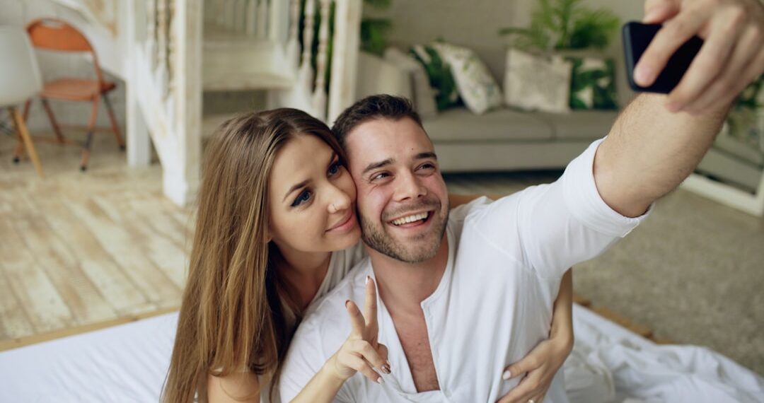 Young beautiful and loving couple take selfie picture on smartphone camera while lying in bed at morning