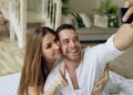 Young beautiful and loving couple take selfie picture on smartphone camera while lying in bed at morning